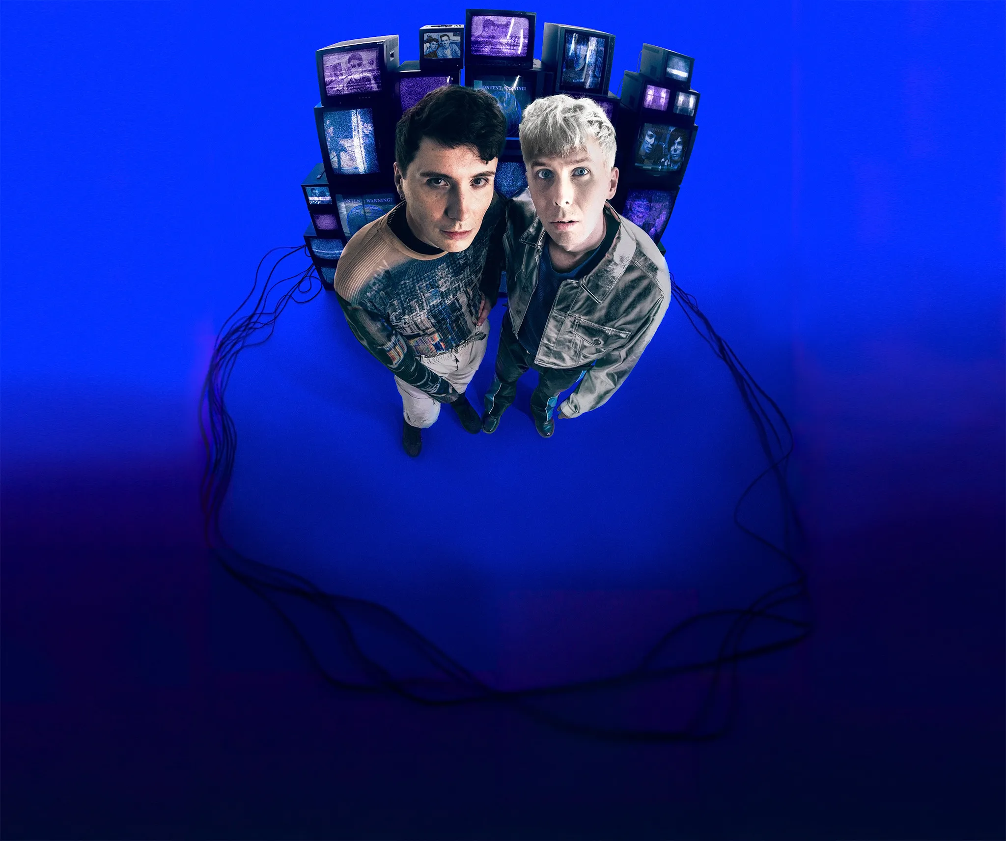 Dan and Phil are stood in front of a stack of old analogue TVs displaying videos from their channel. The image is shot with a fisheye lens and they are styled in late-80s clothing.