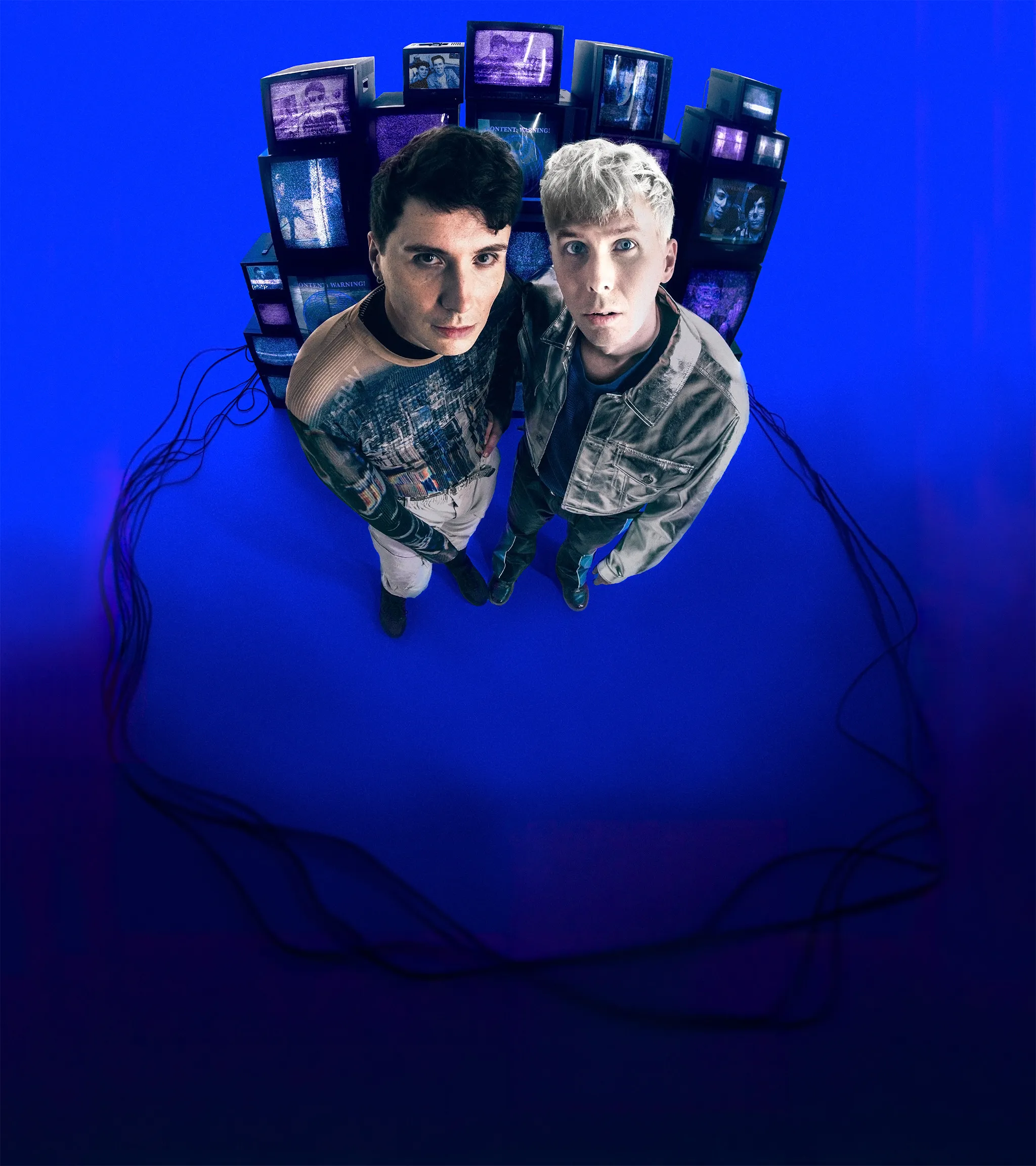 Dan and Phil are stood in front of a stack of old analogue TVs displaying videos from their channel. The image is shot with a fisheye lens and they are styled in late-80s clothing.